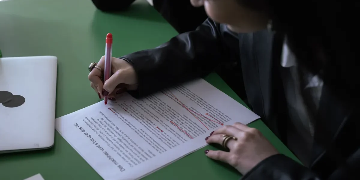 Person proofreading a printed article with a red pen beside a laptop