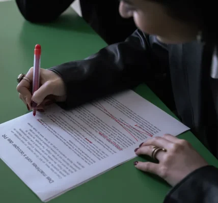 Person proofreading a printed article with a red pen beside a laptop